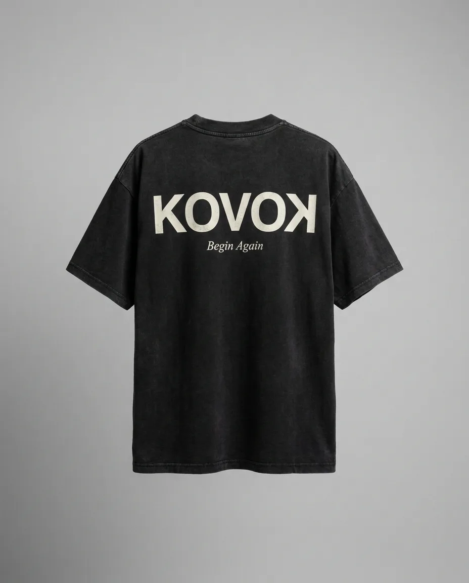 Back of the washed-black tee — large cream KOVOK wordmark with reversed final K above the words Begin Again